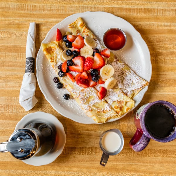Overhead view of crepes topped with strawberries, blueberries, bananas, and powdered sugar, with coffee and syrup.