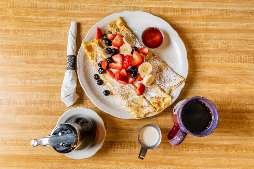 Overhead view of crepes topped with strawberries, blueberries, bananas, and powdered sugar, with coffee and syrup.