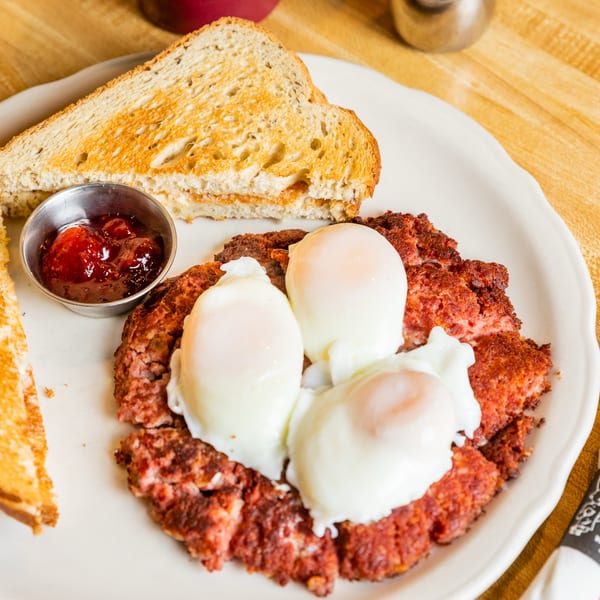 A breakfast plate with corned beef hash, three poached eggs, two slices of toast, and jam.