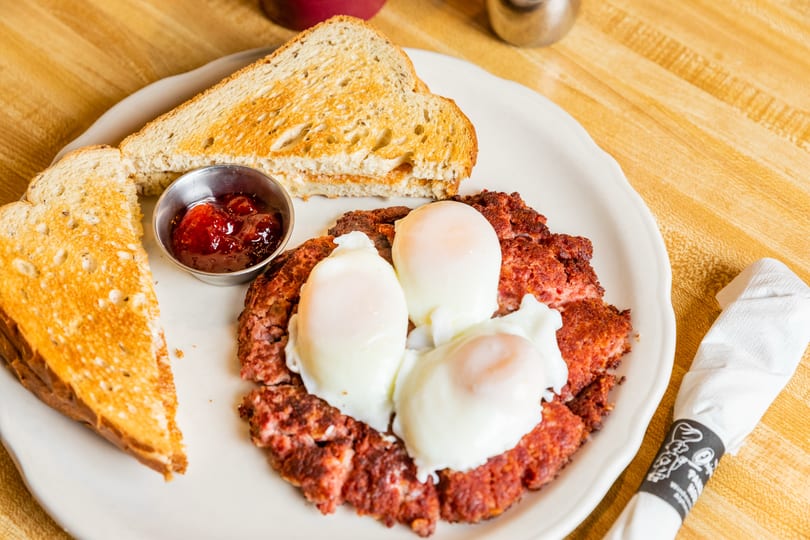 A breakfast plate with corned beef hash, three poached eggs, two slices of toast, and jam.