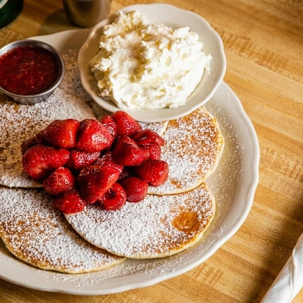Pancakes with powdered sugar, strawberries, strawberry sauce, and whipped cream on a wooden table.