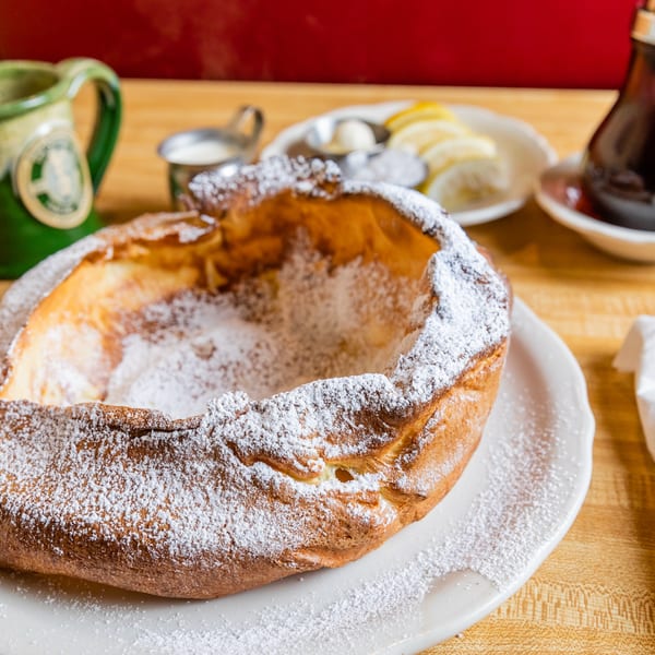 A large, golden-brown Dutch baby pancake dusted with powdered sugar, served with lemon, butter, and syrup.
