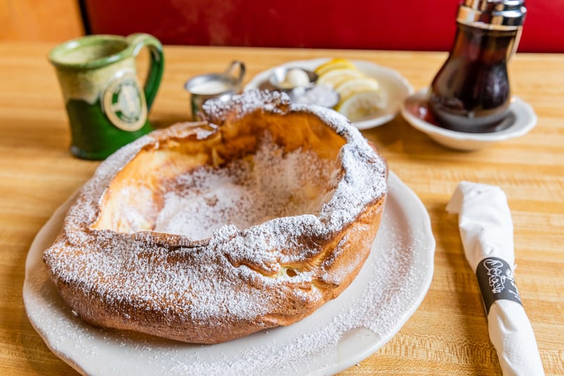 A large, golden-brown Dutch baby pancake dusted with powdered sugar, served with lemon, butter, and syrup.